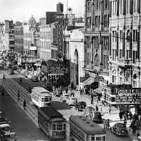 A black-and-white photo of a busy street in Harlem during the Harlem Renaissance period. The photo’s perspective is from a high point directed across the street to capture trolleys and pedestrians.