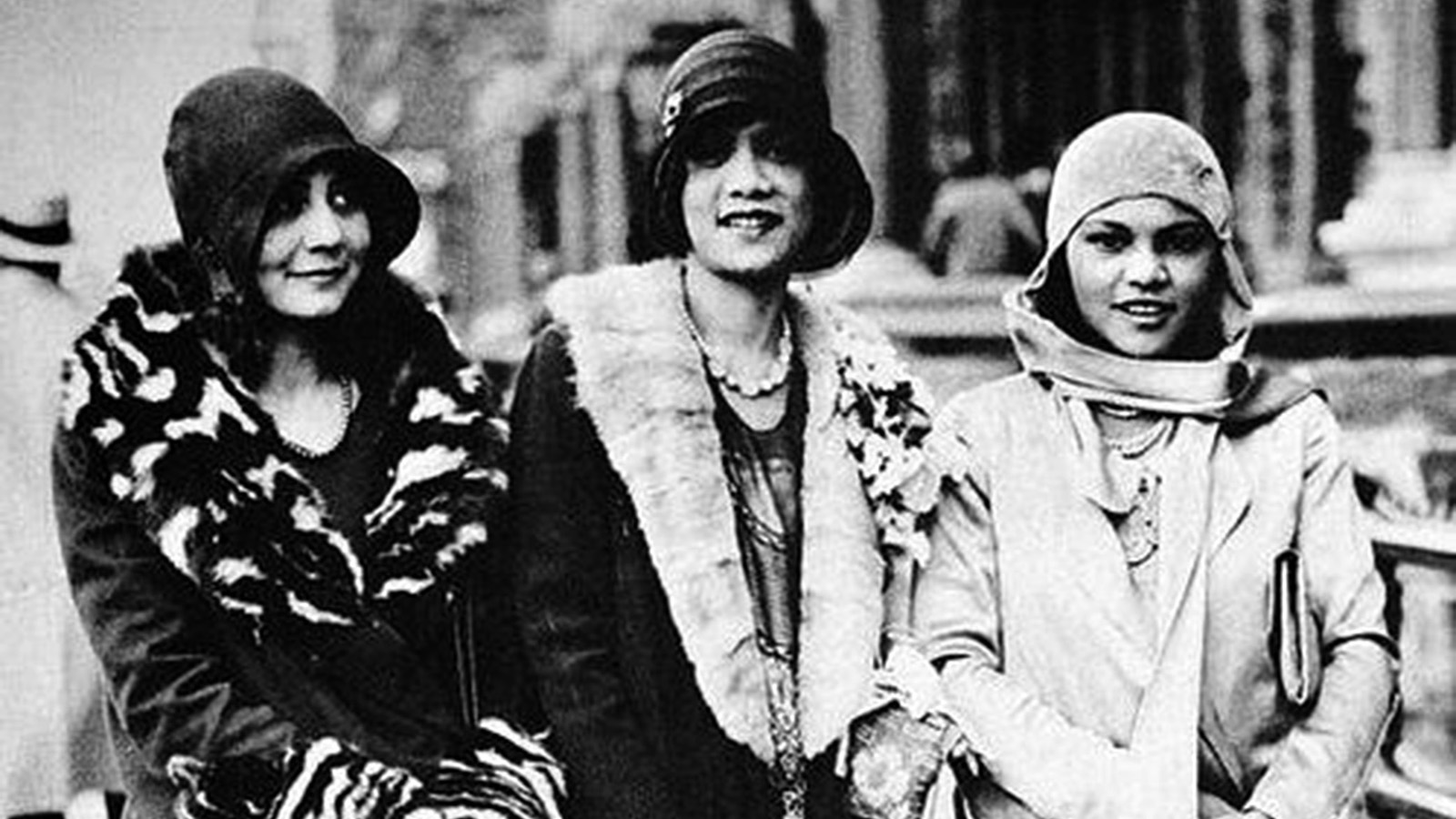 A black-and-white photo of three fashionable women posed in front of a building. Each woman wears a 1920s or 1930s bucket-styled hat that matches her coat.