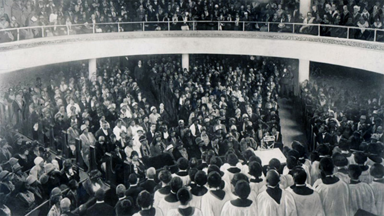 A black-and-white interior photo of a large church congregation sitting in pews on a main floor level and an upper level. A large choir wearing white robes stands in front of the congregation, the singers’ backs to the camera.  