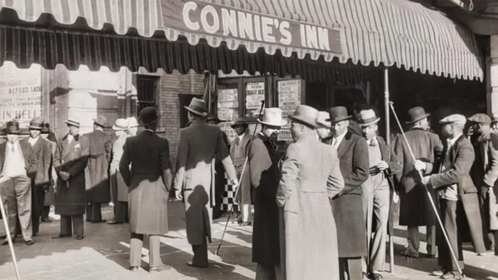 A black-and-white photo of a group of Black men in long dress coats, suits, and top hats waiting outside of a building on which the sign “Connie’s Inn” is displayed. 