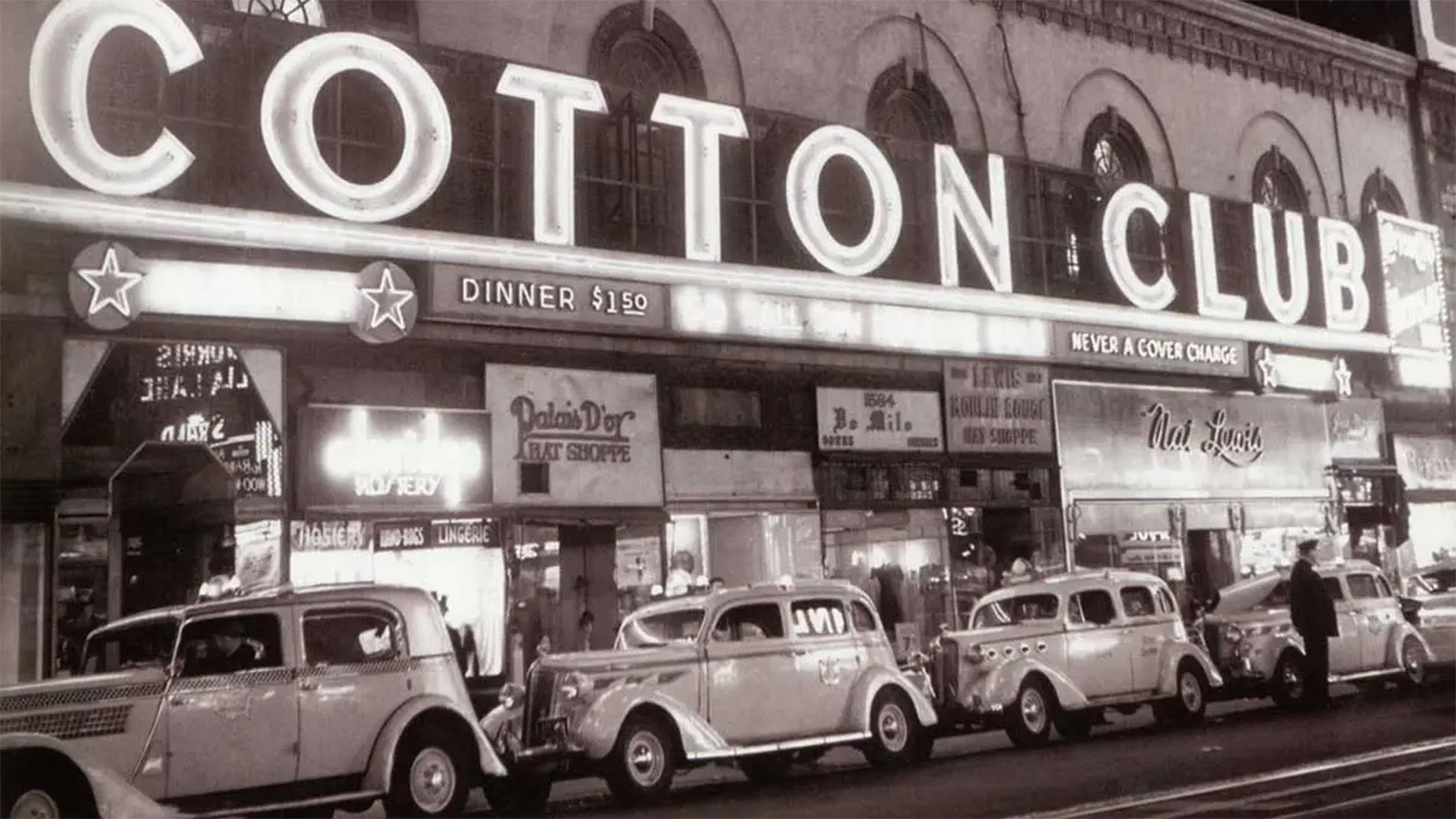 A photo of a street view of the Cotton Club, featuring its main building sign lit up. Old-fashioned vehicles are parked in a line along the street.  