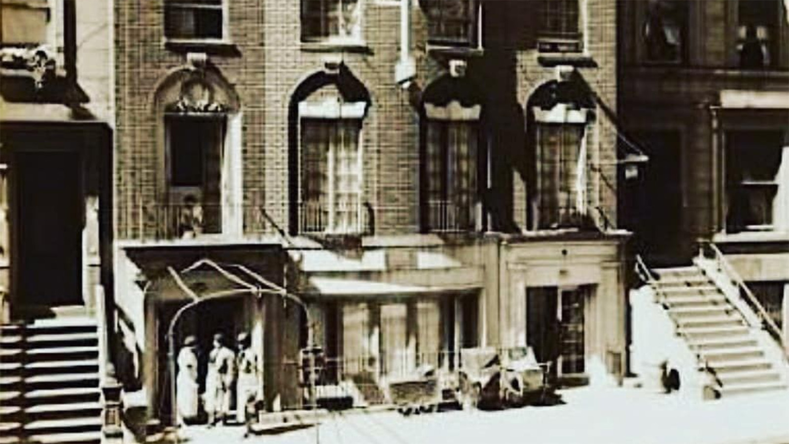 A sepia-toned photo of the front of a large, brick, multi-storied residential building along a street. Three people stand clustered together in one nook on the ground-level.