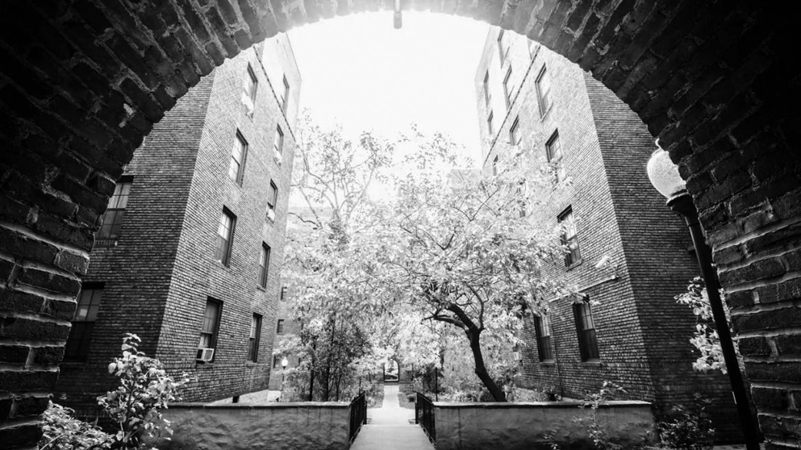A black-and-white photo taken from inside a brick archway entrance that looks out into a small courtyard surrounded on either side by brick apartment buildings.