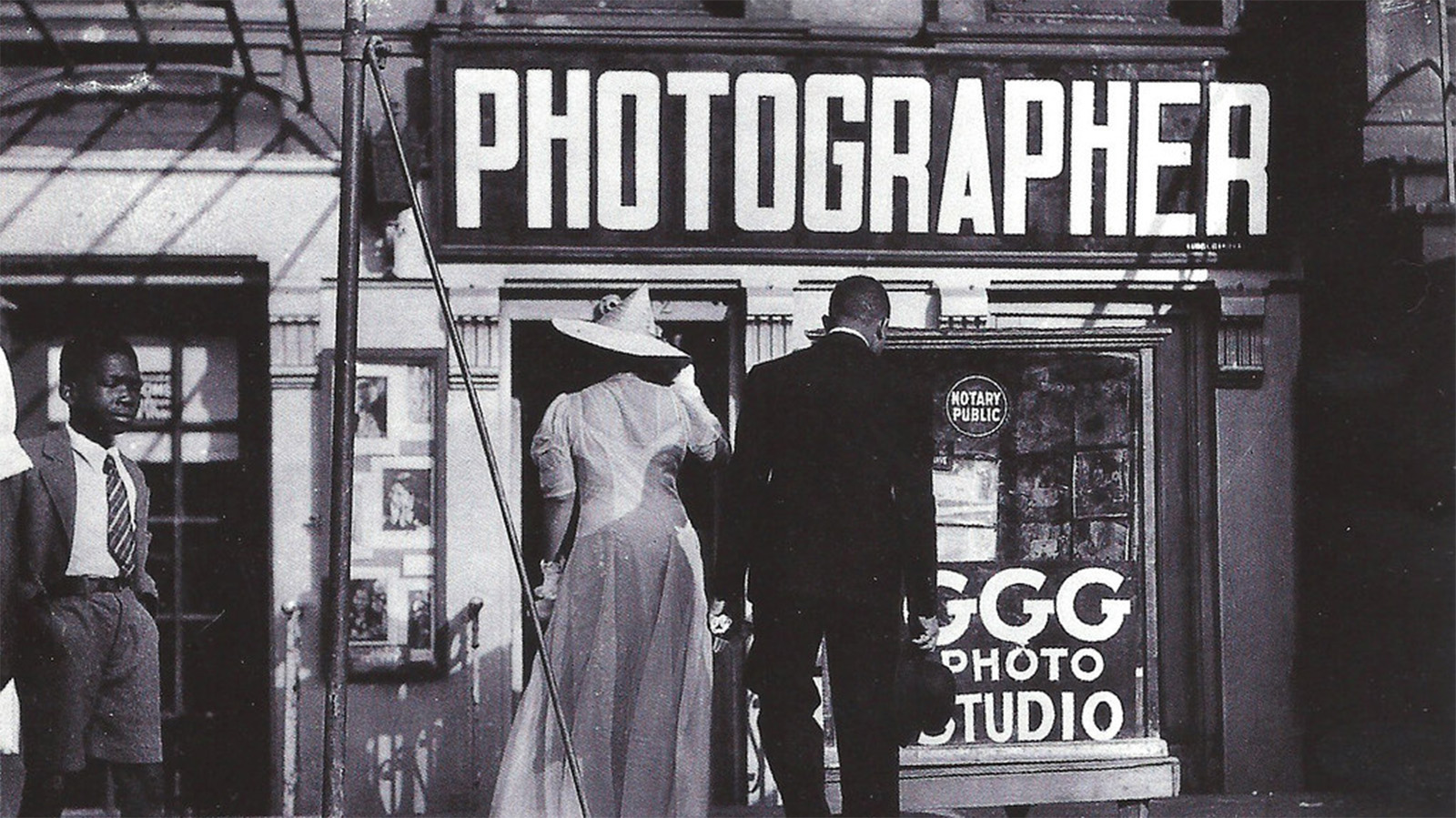 A black-and-white photo of three Black individuals standing outside of a storefront that has one large “PHOTOGRAPHER” sign above its windows and a sign that says “GGG PHOTO STUDIO” on the bottom of the window. The two men in the photo wear business suits while the woman wears a long dress with gloves and a matching pointy hat. The woman and one of the men look at the photography studio’s window displays. 