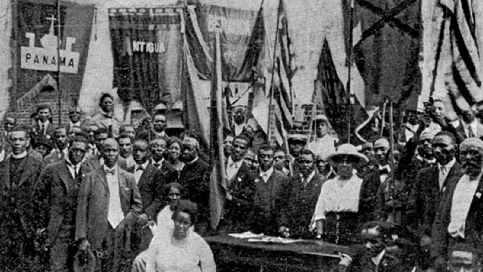 A black-and-white photo of a group of Black men and women dressed in business suits and dresses posing for the camera. Some of the crowd stands around a desk on which a few pieces of paper are visible. Various flags hang from standing poles and the walls around the crowded room. 