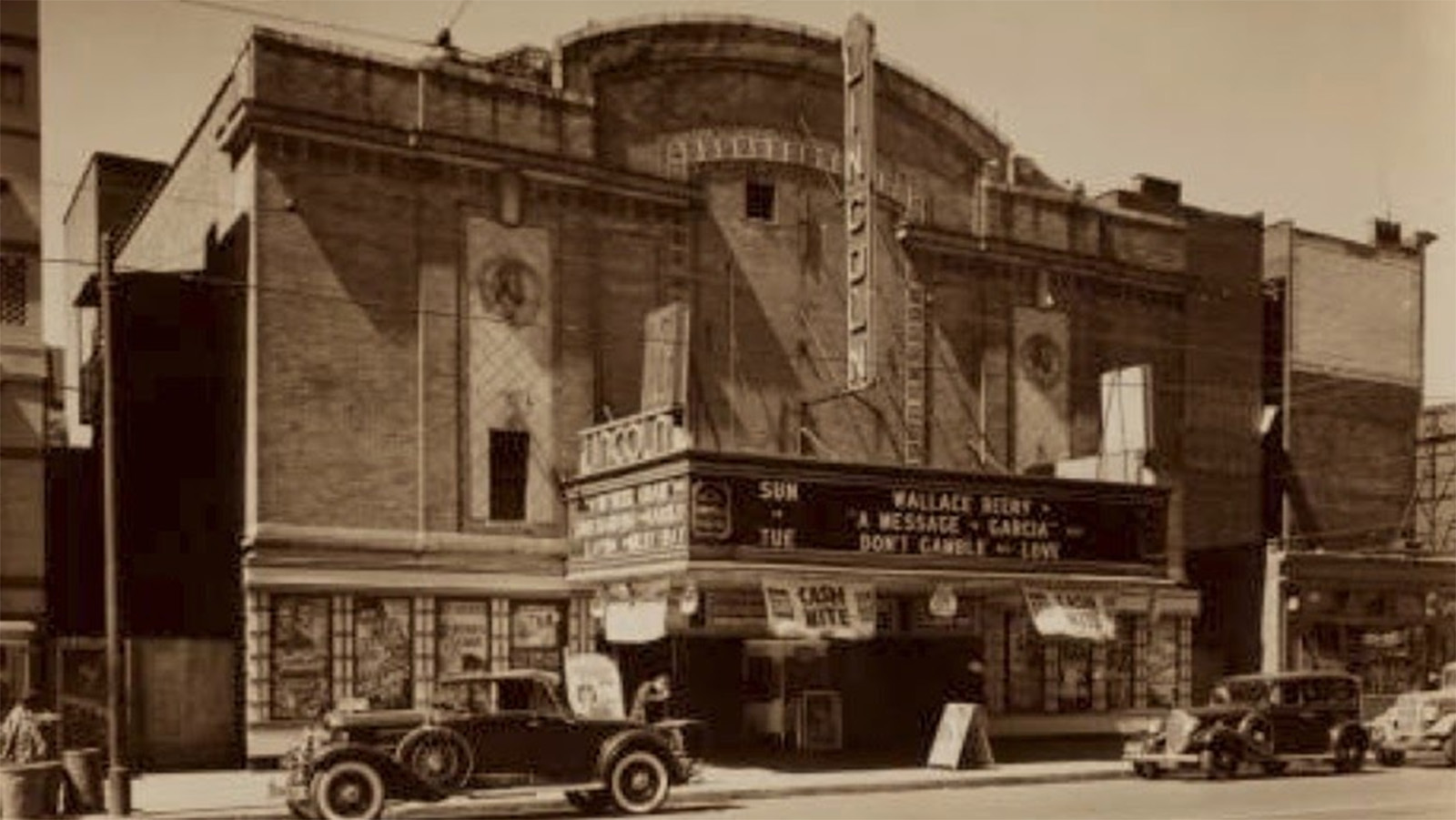 A sepia-toned daytime photo of the front of the Lincoln Theatre in New York. Old-fashioned vehicles are parked on the street in front of the building.
