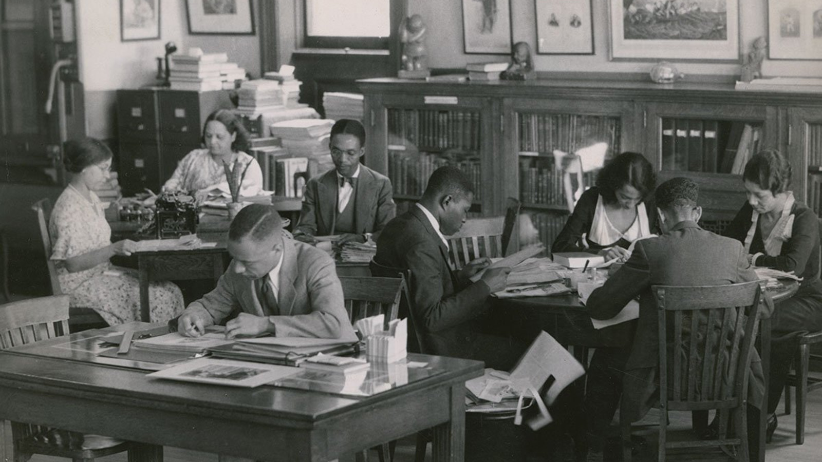A black-and-white photo of Black men and women sitting at three tables in a library setting. Each person is focused on either reading a book or a document or on writing. The background includes bookshelves with small art objects on their surfaces, filing cabinets with stacks of books and binders on their surfaces, and images hanging on the wall. 