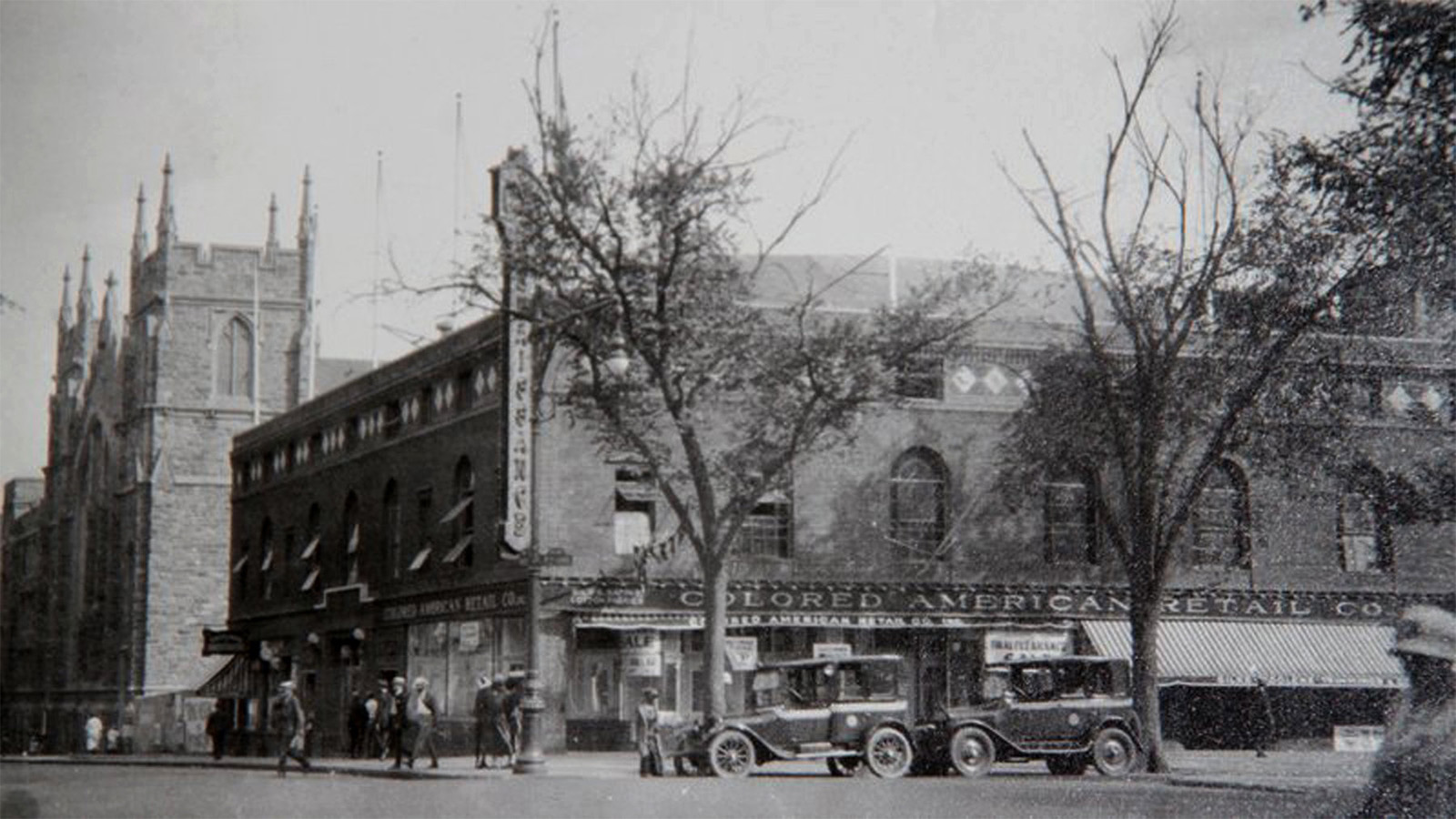    A black-and-white exterior photo of a large brick building on the corner of a street. Two old-fashioned vehicles are parked on the corner. Several pedestrians walk along the sidewalk next to the building. 