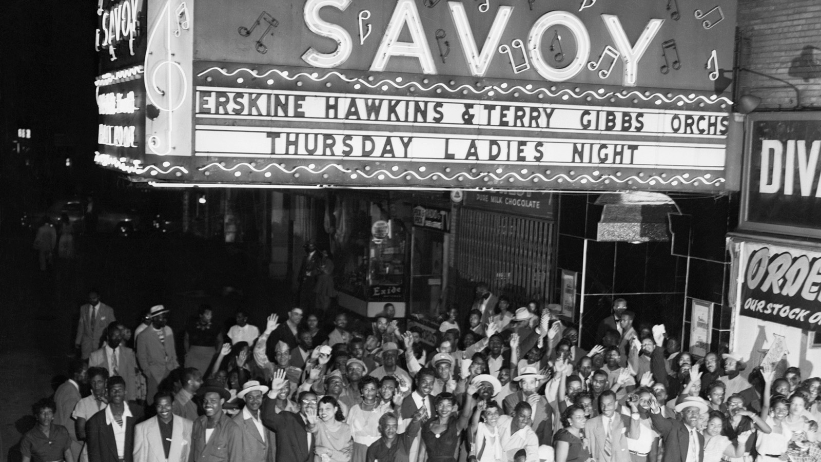 A black-and-white nighttime photo of a large crowd of Black attendees standing underneath the large marquee sign for the Savoy Ballroom. The crowd members are dressed in nice suits and dresses—some hold their hands up in the air in a friendly gesture toward the cameraperson. 