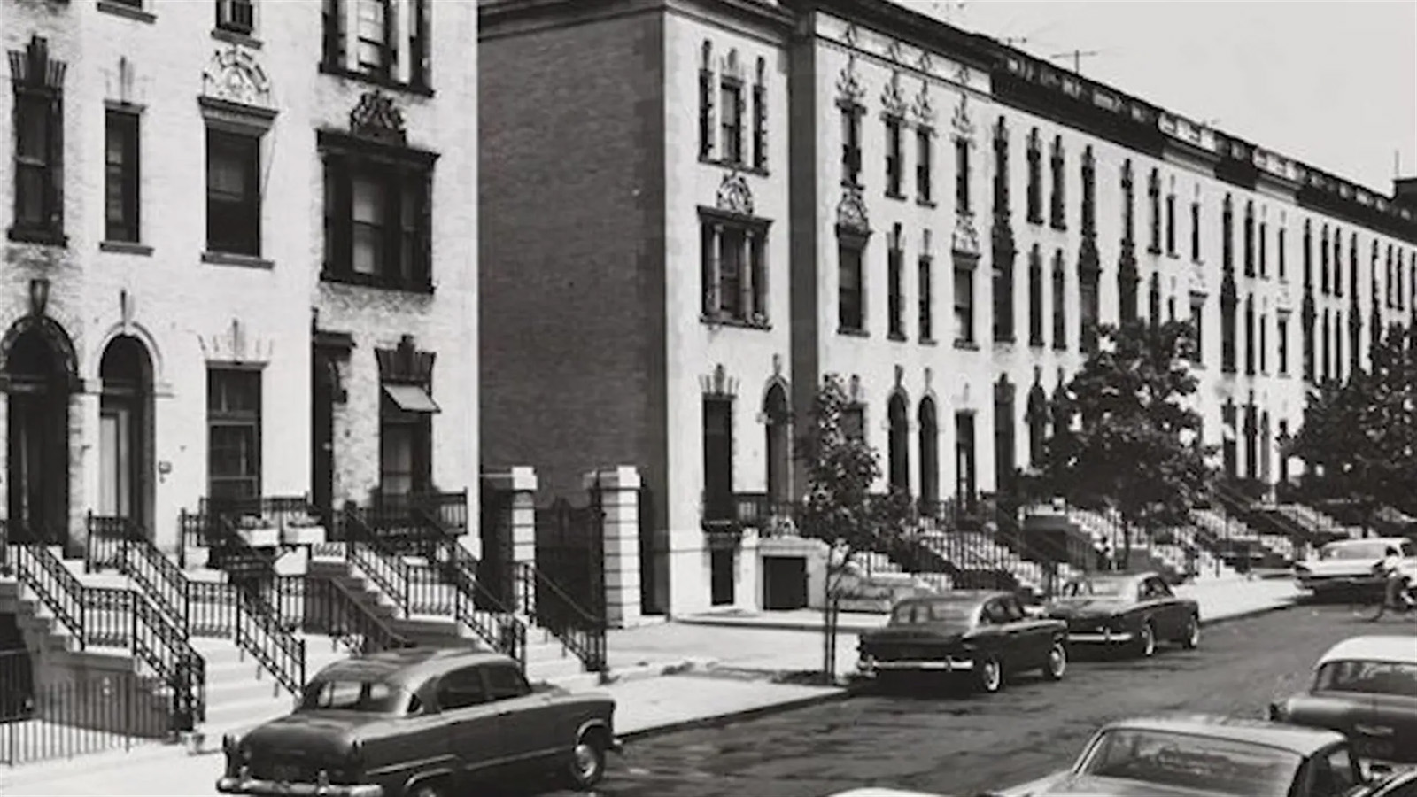 A black-and-white photo of a street of townhouse-styled houses. Several dark-colored old-fashioned vehicles line the street in front of the homes. 