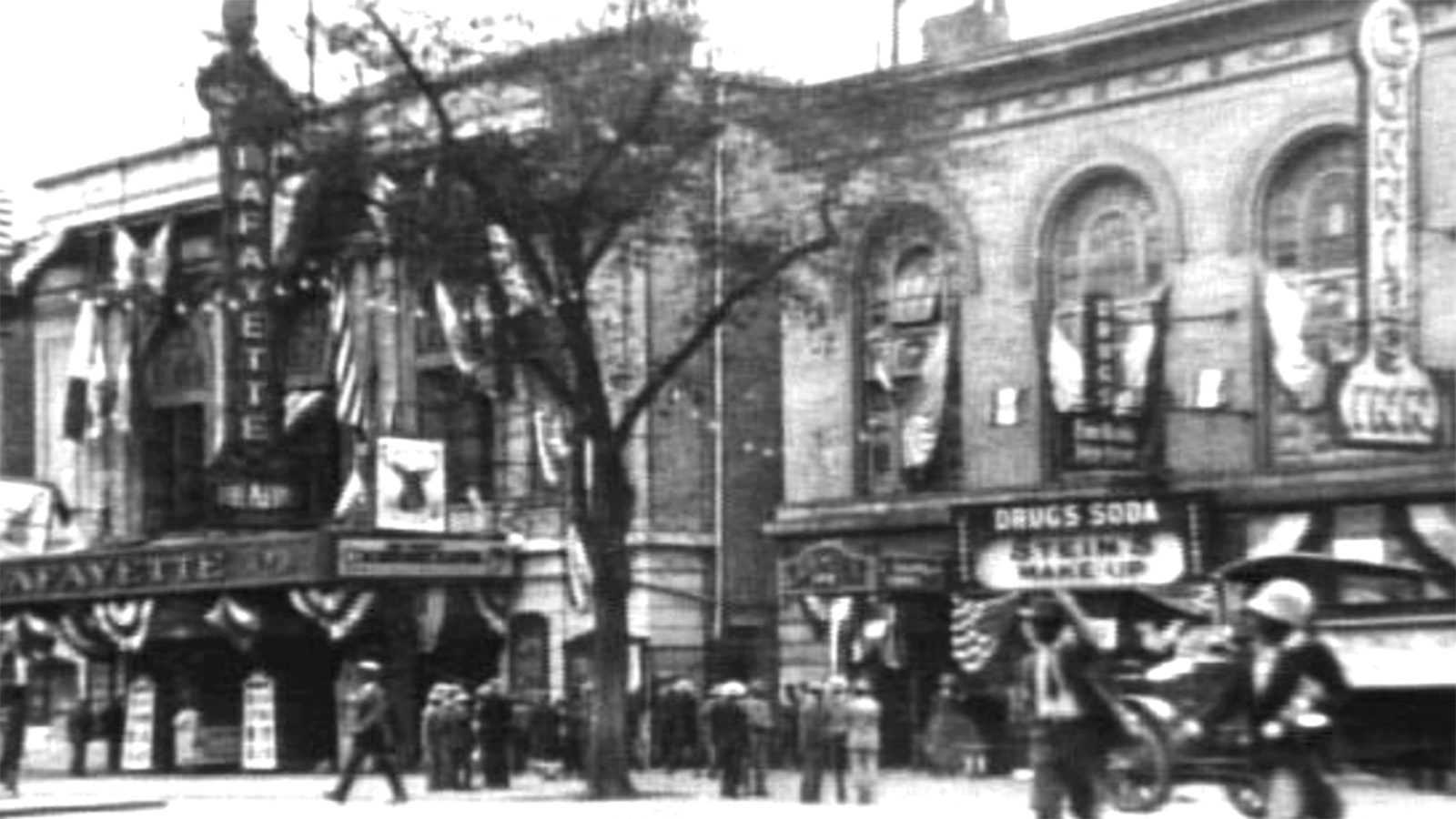 A black-and-white photo of a tall chestnut tree located in front of a building with “Lafayette Theater” signage and a building with “Connie’s Inn” signage on their fronts. Black civilians in business suits congregate in small groups near the tree while others walk along the street.   