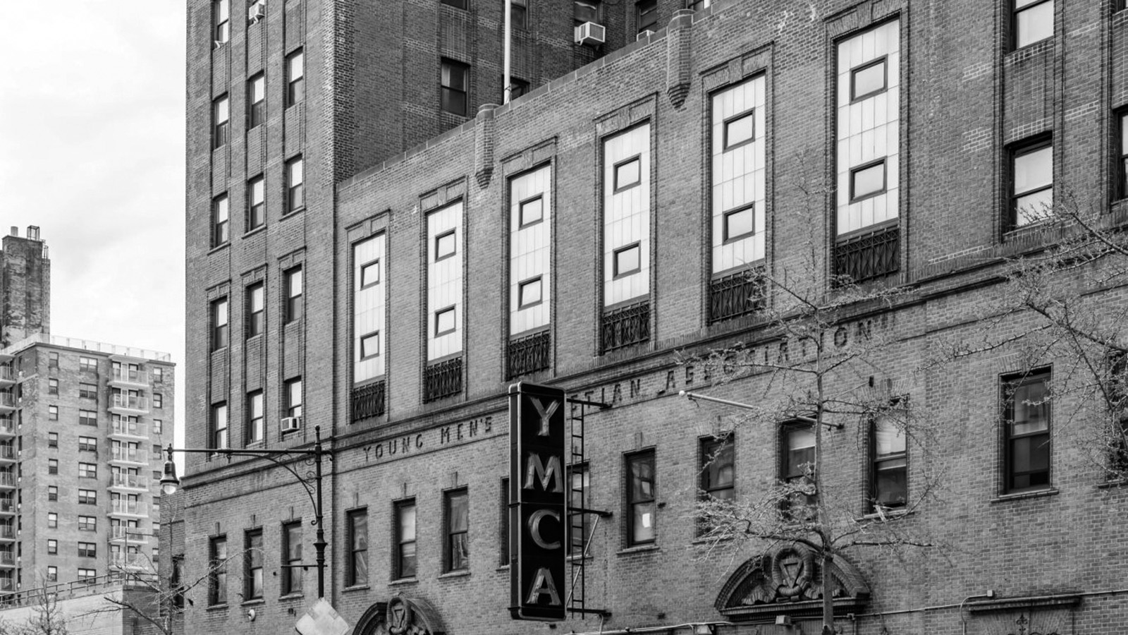 A black-and-white image of the front of the Harlem YMCA building, which centers both its vertical “YMCA” sign and the “Young Men’s Christian Association” engraving on the building.  