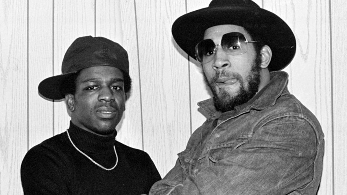 Black and white photo of hip hop pioneers DJ Tony Tone wearing a black baseball cap turned to the side, a black turtleneck sweater, and gold chain and DJ Kool Herc wearing a black fedora, denim jacket, and sunglasses.