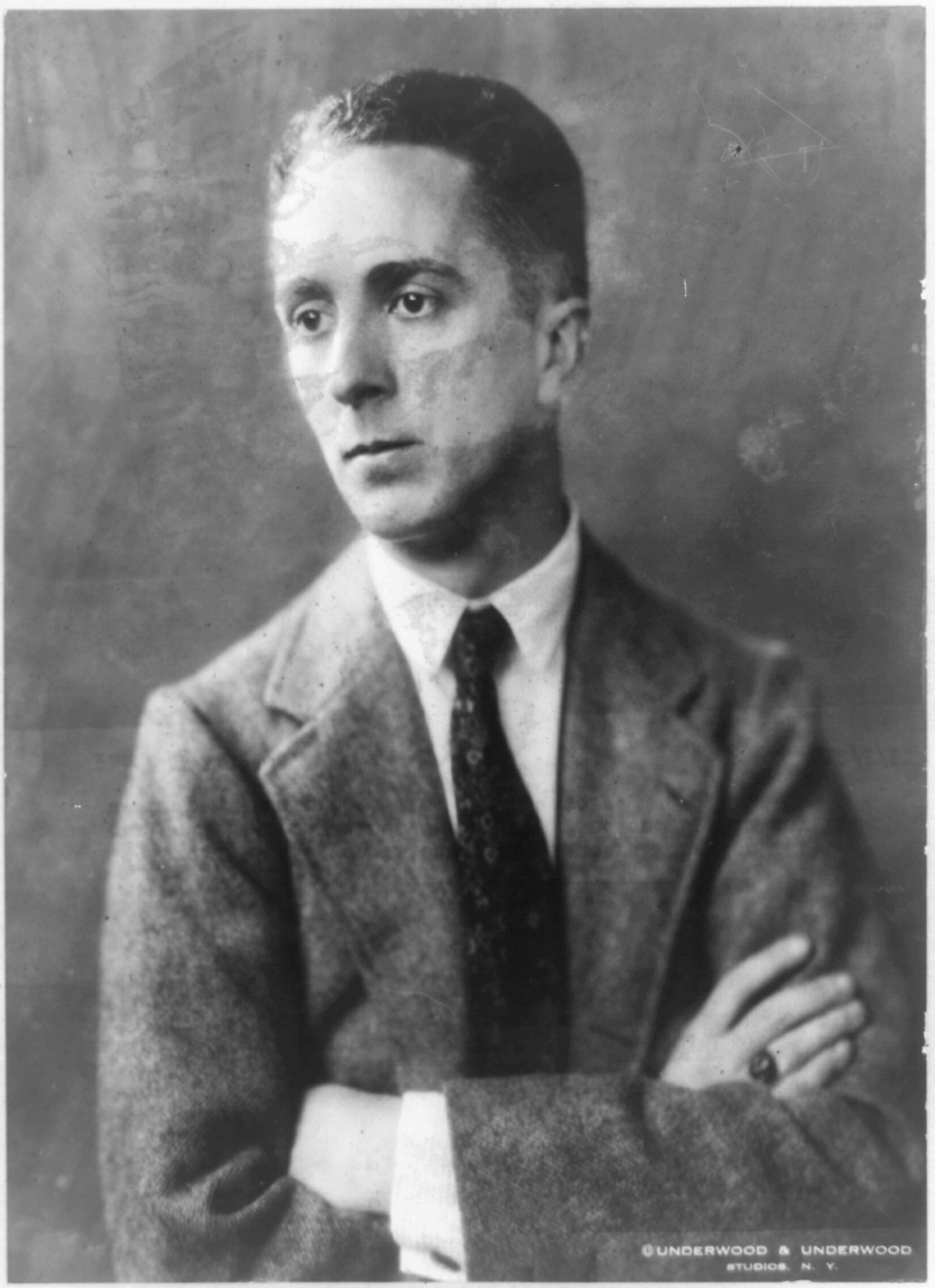 In a vintage photo of artist Norman Rockwell, the man wears a dark jacket with a white dress shirt and dark tie. His arms are crossed at his chest as he looks off to the side.