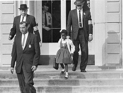 In a black and white photo, a little Black girl (Ruby Bridges) walks between three white adult men as they descend steps outside of a building. She carries a school bag in one hand. The men all have badges on their jackets, and two of the men wear matching arm bands.