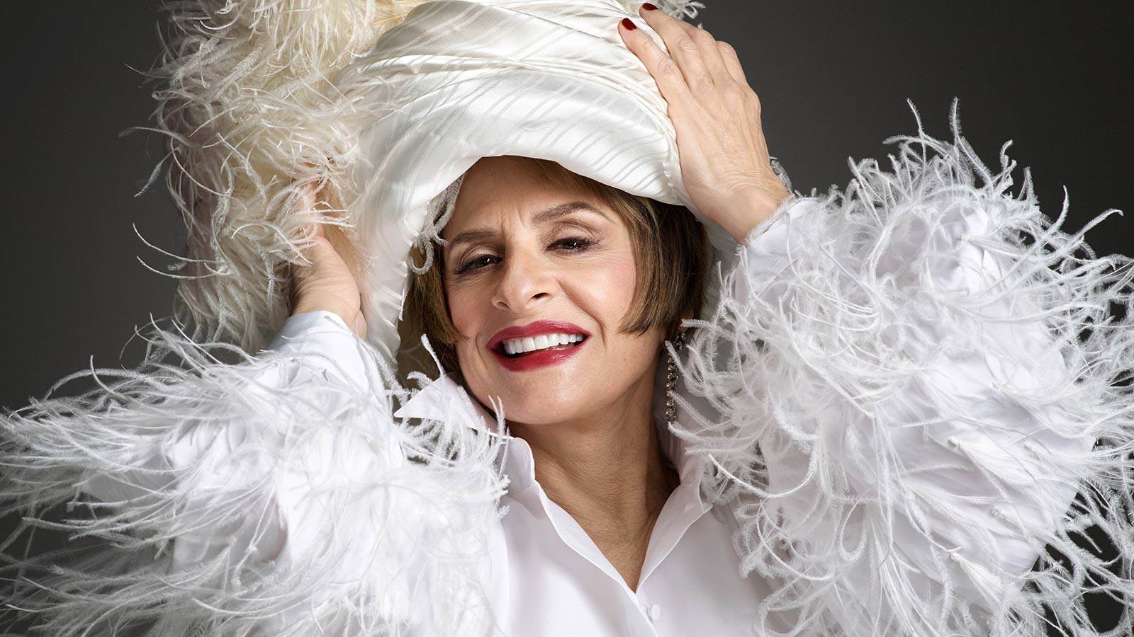 Patti Lupone in white outfits surrounded with feathers on sleeves and hat